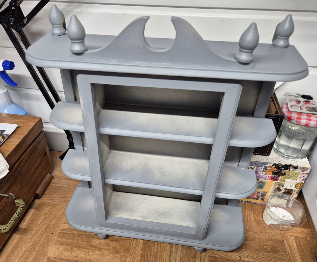 A gray wooden shelf unit featuring three open shelves and decorative finials on top, placed in a room with various items in the background.