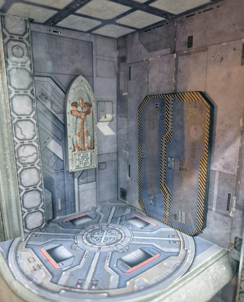 Interior view of a futuristic sci-fi themed room featuring metallic walls, a circular patterned floor, and various technological details including a wall-mounted object and a door with caution stripes.