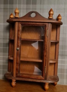 A small wooden display cabinet with a glass door and multiple shelves, featuring decorative finials on top.