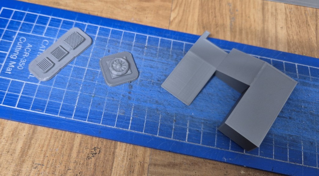 Gray 3D printed components on a blue cutting mat, including two detailed panels and an L-shaped piece.