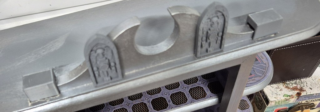 Close-up of a metallic shelf featuring decorative elements, including arched designs and carved figures, set against a gray background.