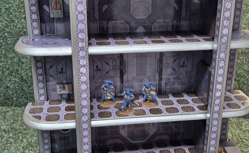 Three miniature figures in blue armor standing on a multi-tiered sci-fi terrain piece with visible metallic floors and walls.
